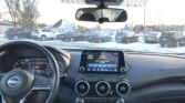 Interior of a car cockpit viewed from the driver’s seat; steering wheel on the left, central touchscreen display, and round air vents visible, with a snowy parking lot outside.