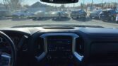 View from a car's front seats: dashboard and rearview mirror, with a busy parking lot and storefronts visible through the windshield on a sunny day.