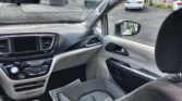View of a car interior from the back seat: dashboard with a central touchscreen, climate controls, gear selector, and light-colored trim; passenger door and side mirror visible.