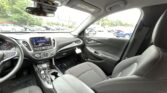 Interior of a modern car cabin seen from the front passenger seat: dashboard with touchscreen, center console, gear shifter, steering wheel, and front seats; exterior parking lot visible through windows.