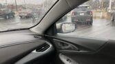 View from inside a car on a rainy day, raindrops on the windshield with a parking lot and cars visible ahead and to the side.