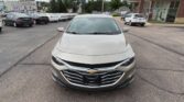 Front view of a beige Chevrolet sedan parked in a car dealership lot with other cars in background.