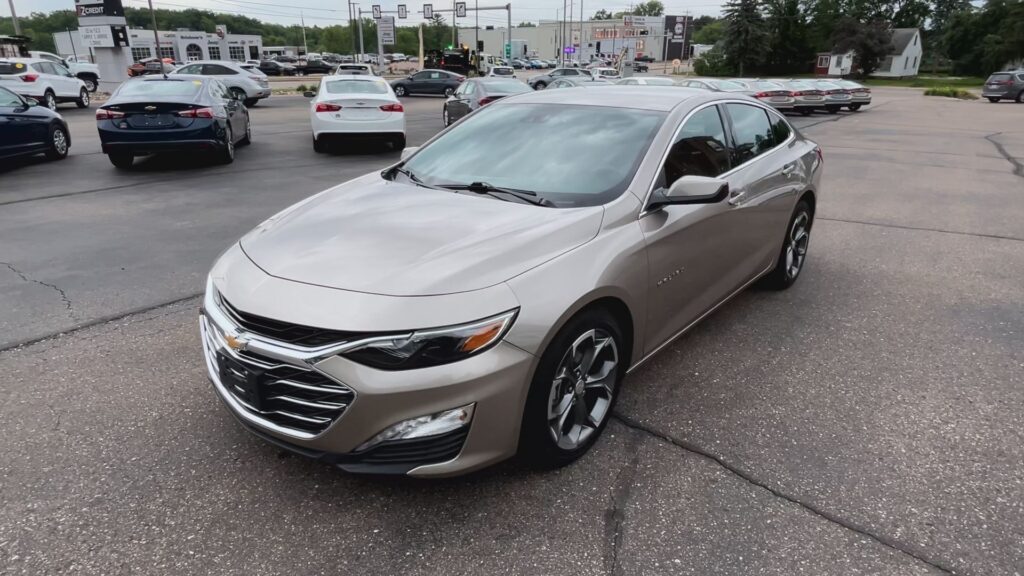 Front three-quarter view of a silver Chevrolet Malibu in a busy car dealership lot with other cars and a building in the background