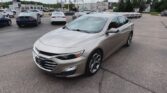 Front three-quarter view of a silver Chevrolet Malibu in a busy car dealership lot with other cars and a building in the background