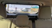 Back seat view of a car, looking through the rear window at a dealership lot with a green-roofed building and parked cars behind.