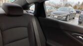 Rear passenger seat and headrest with dark fabric upholstery; view through the window shows a gray Chevrolet sedan parked in a parking lot on a cloudy day, with other cars nearby.]