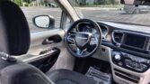 Interior view of a car cockpit: steering wheel with Chrysler logo, instrument cluster, center touchscreen, and drive-side door panel with window controls.