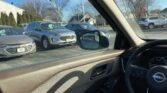 View from inside a car looking out the driver-side window at a parking lot with silver SUVs, leafless trees, and a bright blue sky; side mirror in foreground.