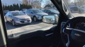 Interior view from a car looking out at a dealership lot; a silver sedan is in the foreground with other cars and a white building in the background.