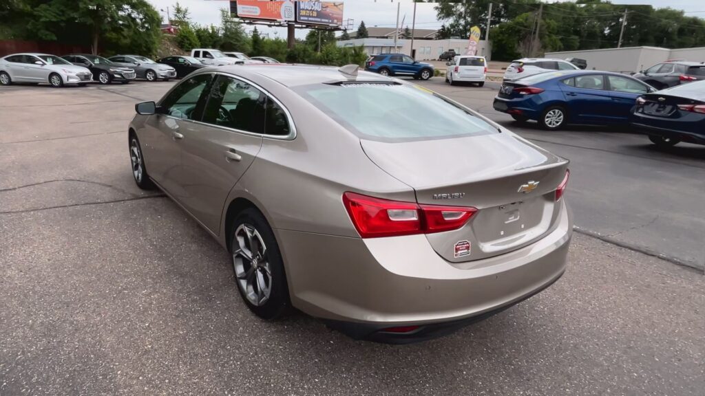 Rear three-quarter view of a beige Chevrolet Malibu in a car dealership lot, with other cars and billboards in the background.