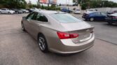 Rear three-quarter view of a beige Chevrolet Malibu in a car dealership lot, with other cars and billboards in the background.