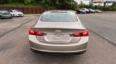 Beige Chevrolet Malibu seen from the rear in a car dealership lot with other cars parked in the background and a wooden fence along the left side.