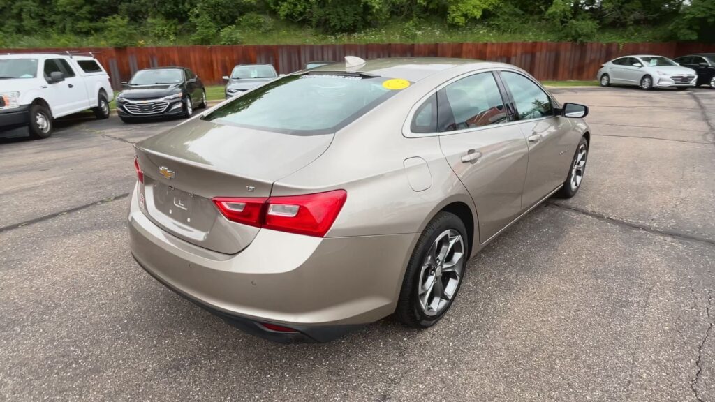 Beige Chevrolet sedan viewed from the rear-left in a parking lot with other cars and a wooden fence behind.