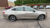 Side view of a silver four-door sedan parked in a car lot with other cars and a wooden fence in the background, alloy wheels visible.] ,