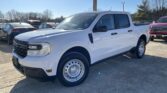 White Ford pickup (F-Series) shown from a front-left angle in a car lot, with other vehicles and a gravel lot under a clear blue sky.