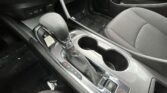 Interior of a car center console with the gear selector, two cup holders, and electronic parking brake; a small storage area behind the shifter contains a plastic cover and papers on the floor mat.