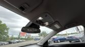 Car interior ceiling with illuminated overhead console and rearview mirror, viewed from the front seat in a parked car outside a lot activity.