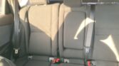 Rear car seat view showing three seating positions with belts and buckles, sunlit fabric upholstery, headrests visible on the outer seats.