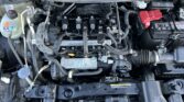 Under-hood view of a car engine bay showing the battery on the right, coolant reservoir on the left, and multiple hoses and wiring connecting to the engine block in the center.