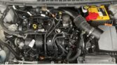 Under-hood view of a vehicle showing the engine, wiring harnesses, and a bright yellow car battery with a red positive cover on the right side; air intake tube and various hoses are visible across the bay.
