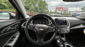 View of a car interior from the driver seat: steering wheel with Chevrolet emblem, instrument cluster, center touchscreen, gear shifter, and door controls; outside are parked cars and billboards.
