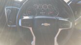 Driver's-eye view of a car dashboard with a circular steering wheel bearing a Chevrolet logo, and an instrument cluster including speedometer, tachometer, and gauges, with sun glare across the windshield.