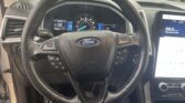 Close-up of a Ford steering wheel with instrument cluster and a right-side touchscreen displaying 106.9 FM radio. Outside cars visible through the windshield.