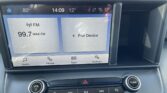 Car infotainment touchscreen showing FM radio (99.7 MAX FM) and a 'Pair Device' tile; bottom row shows Audio, Phone, Apps, Settings with two knobs (Volume and Tune).