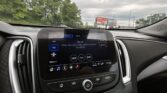 Car dashboard with touchscreen showing FM radio 101.9 and song info, seen through a windshield with trees and billboards outside on a cloudy day
