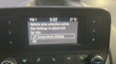 Car stereo display showing FM1 at 5:02 and a message: 'Vehicle data collection active. See Settings to adjust and for info.' with a highlighted 'Connectivity settings' box and OK option nearby. Outside view shows blurred traffic through windshield.
