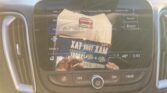 Car infotainment touchscreen displaying radio interface (FM 95.7), with reflection of a magazine and hands in view; dashboard controls visible.