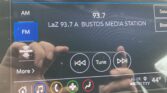 Car infotainment touch screen showing FM radio 93.7 LaZ Bustos Media Station with playback controls (rewind, play, fast‑forward) and a blue FM button.