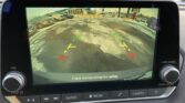 Rearview camera display with parking guidelines (green/yellow/red) over a snowy parking lot; warning text: Check surroundings for safety.