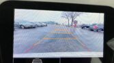 Rear-view camera display showing a parking lot with guiding orange and red lines for backing up, surrounding cars and a tree in the distance.