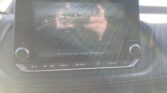 Car dashboard screen showing the rear-view camera feed with guiding lines as the car backs up in a parking lot, text reads 'Check surroundings for safety'