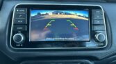 Car's in-dash display shows rearview camera with colored guide lines and a street scene behind the vehicle, warning to check surroundings for safety.