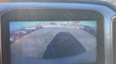 Rearview camera display showing a parking lot with yellow guiding lines and surrounding cars, sun glare on the screen