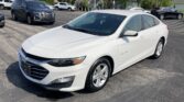 White Chevrolet Malibu in the foreground at a car dealership lot with other vehicles and trees in the background.